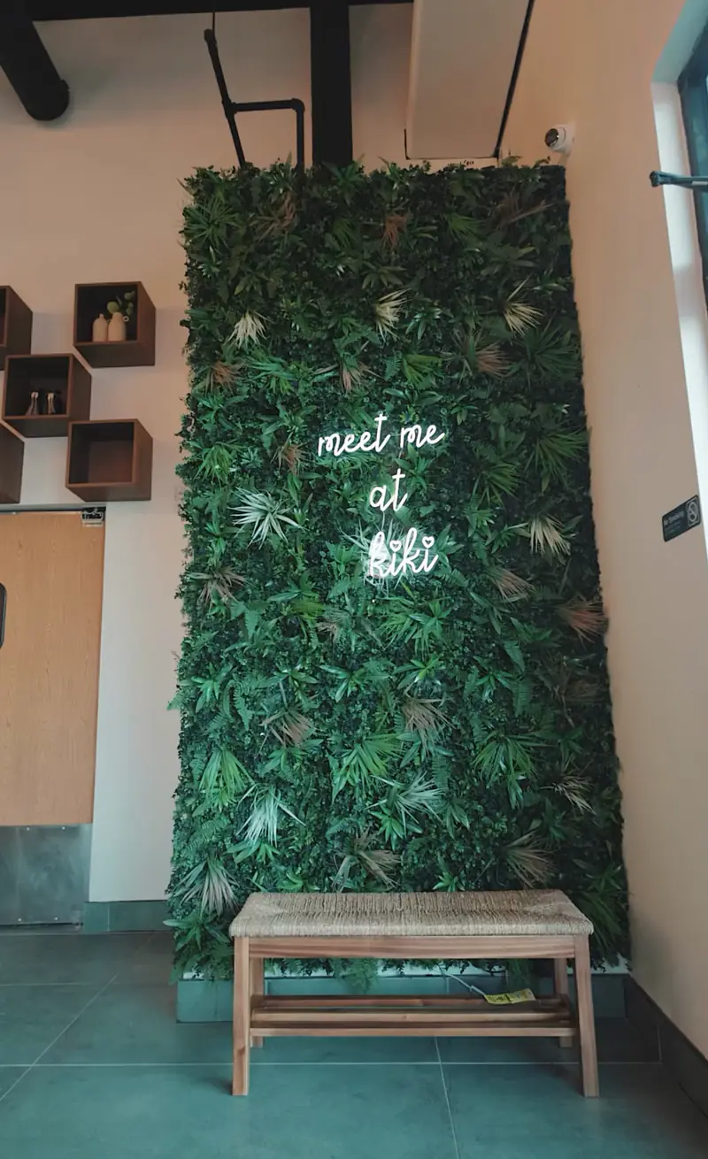 Green plant wall with a "meet me at kiki" neon sign and a bench